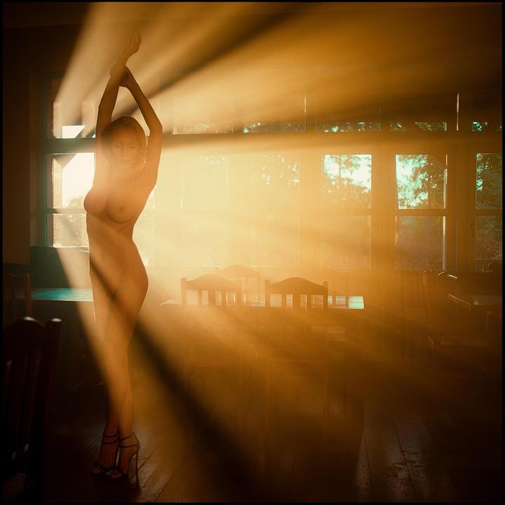 A nude woman stands with her arms raised in a room filled with dramatic light rays.