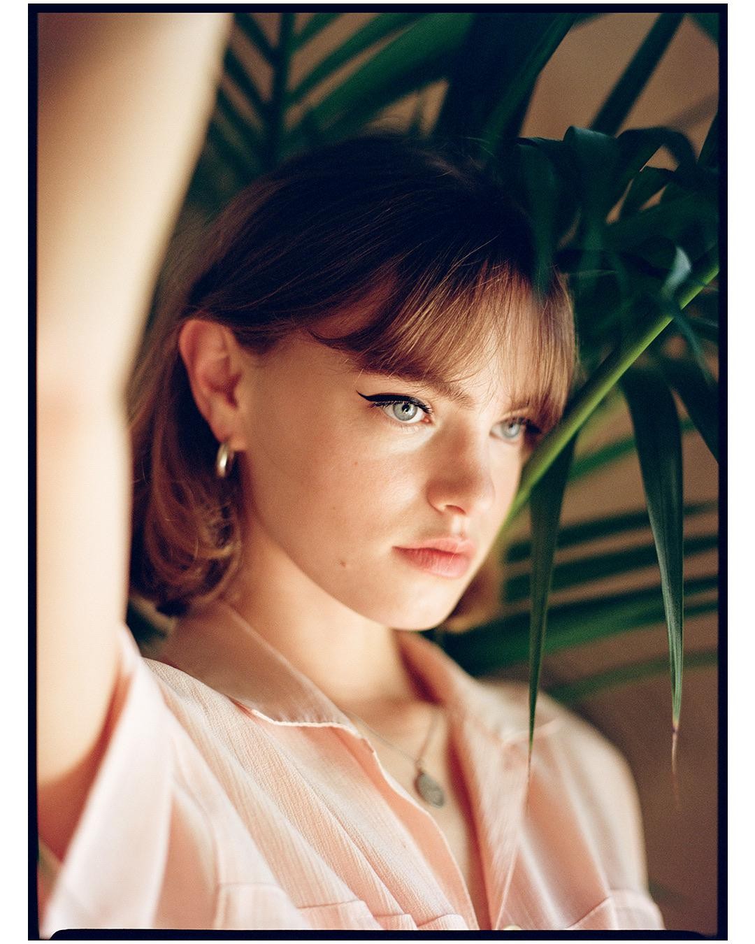 A woman with bangs and eyeliner looks to the side, partially obscured by a large green plant.