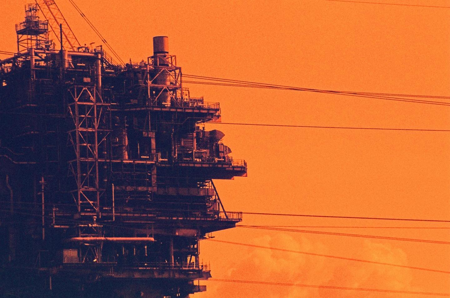 An industrial structure with many pipes and platforms is silhouetted against an orange sky.