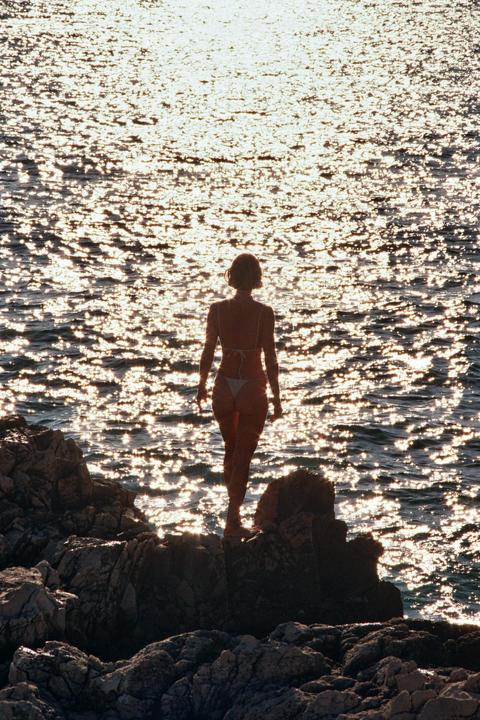 A woman in a bikini stands on rocks looking out at the sparkling water.