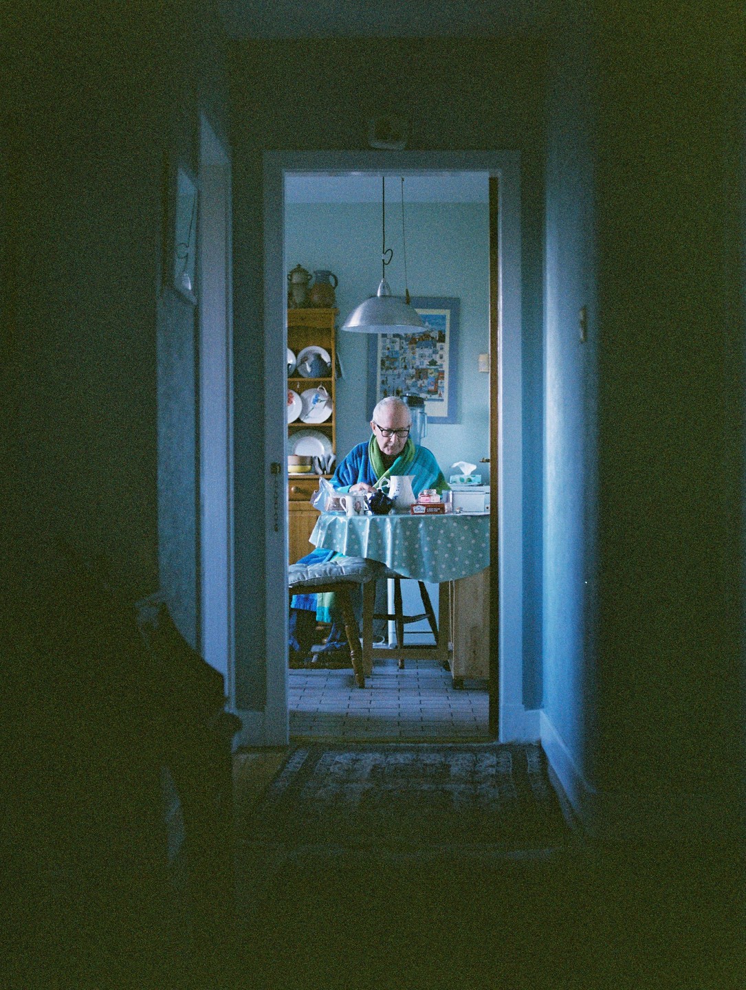 A man in a blue robe and green scarf sits at a table in a kitchen, viewed through a doorway.