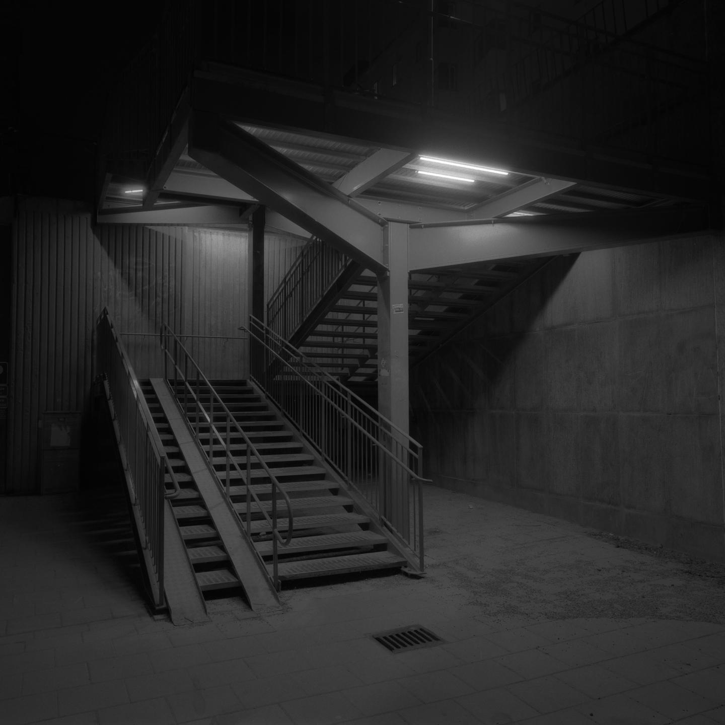 A dark, empty underpass features a set of stairs with a ramp and a second set of stairs leading upwards.
