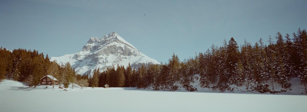 A snow-covered mountain rises behind a frozen lake and a forest with a small cabin.