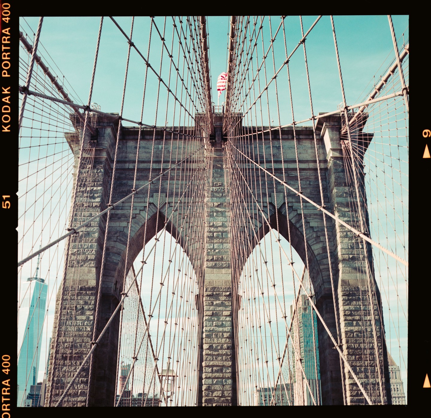 The Brooklyn Bridge is seen from below, with its stone arches and numerous suspension cables visible against a clear sky.