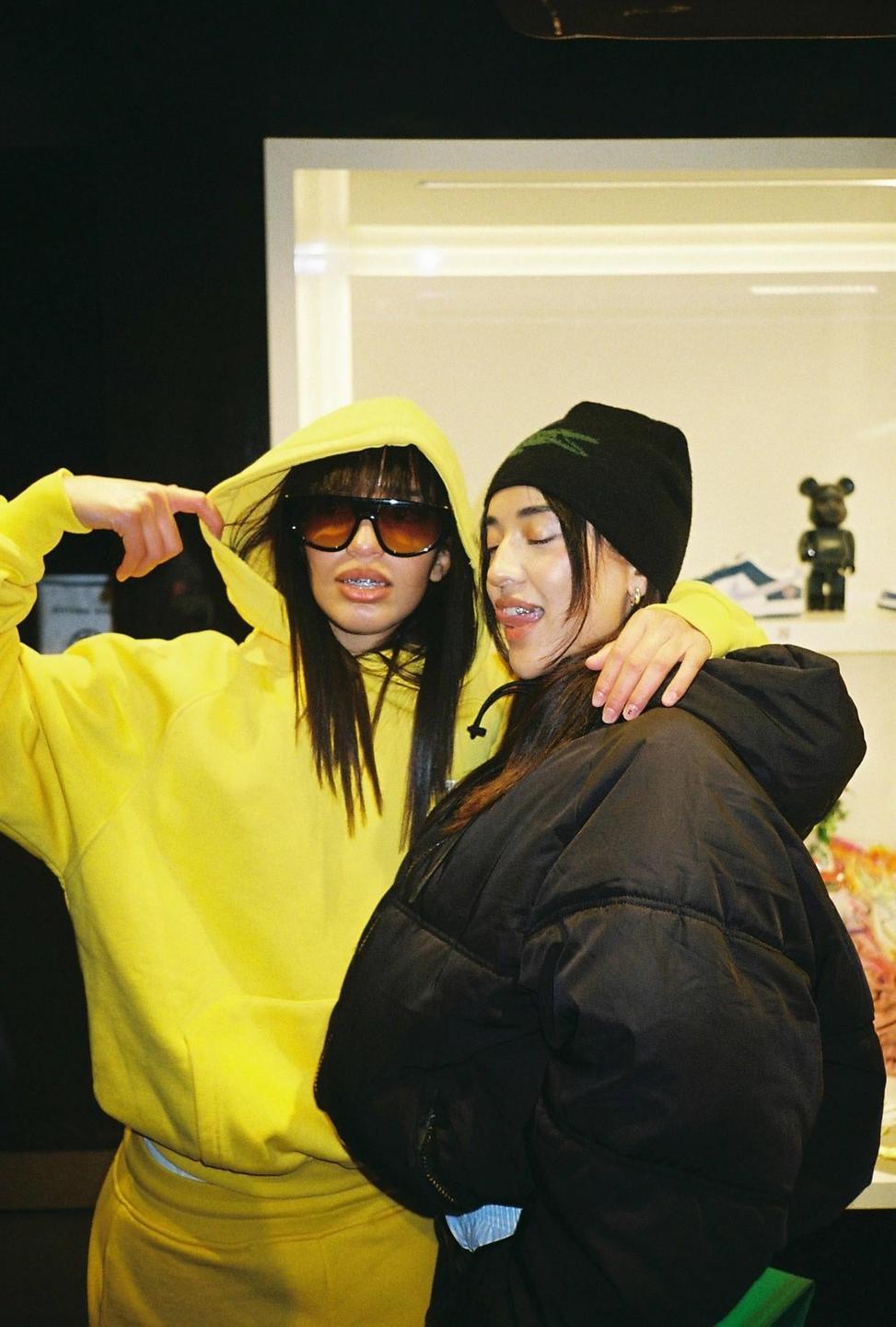 Two women pose together, one in a yellow hoodie and sunglasses, the other in a black puffer jacket and beanie.