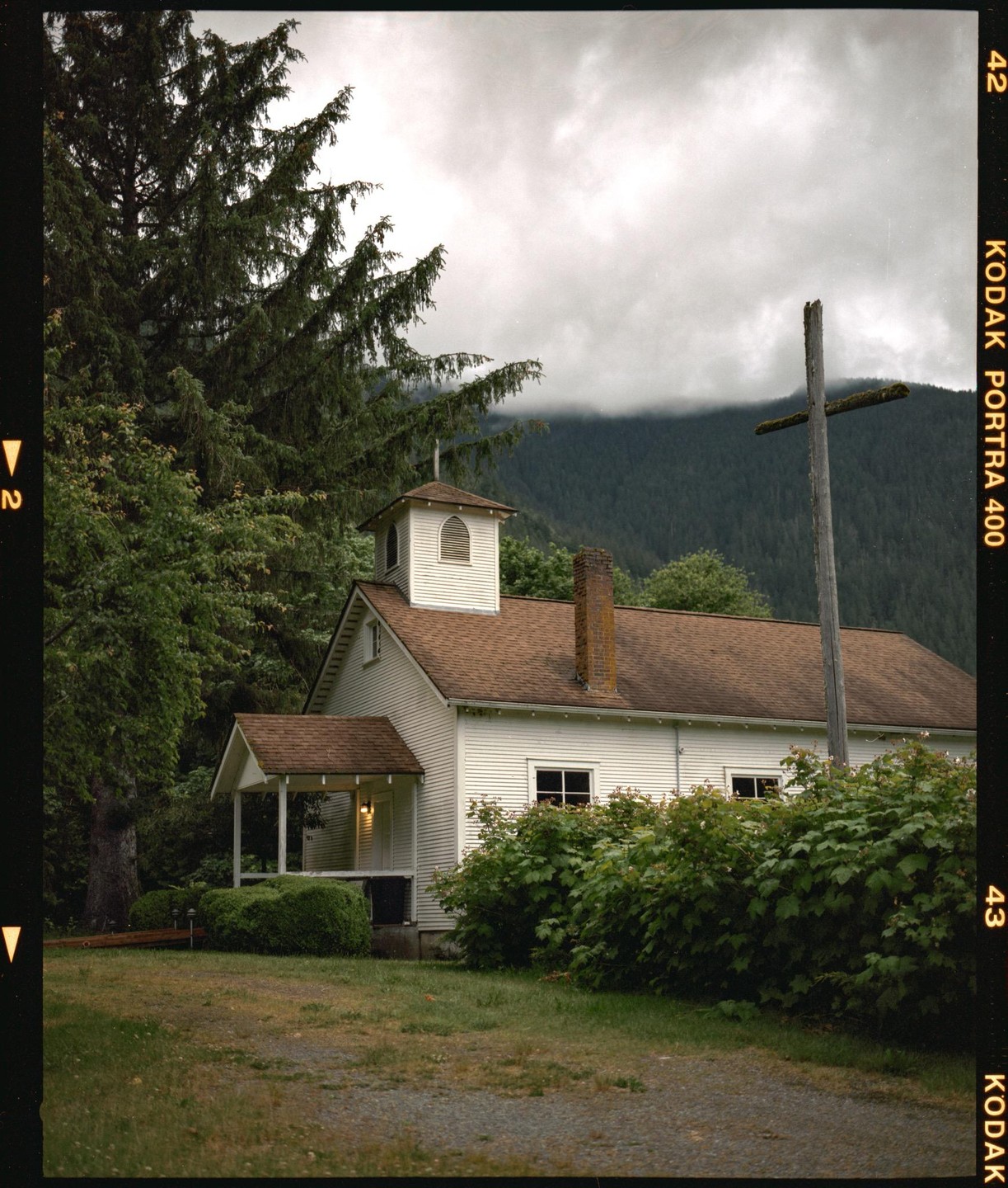 A white church with a brown roof stands next to a large wooden cross, surrounded by trees and mountains under a cloudy sky.