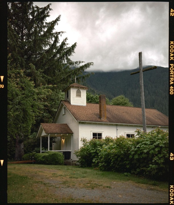 A white church with a brown roof stands next to a large wooden cross, surrounded by trees and mountains under a cloudy sky.