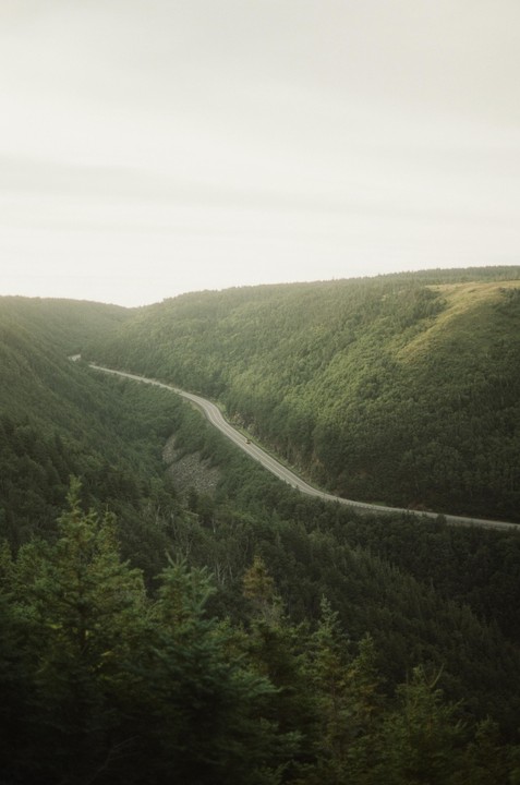 A winding road cuts through a lush, green valley under a bright sky.