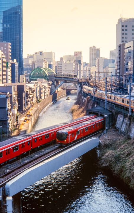 Two red trains are on tracks above a canal with a small boat, surrounded by buildings and other train lines.
