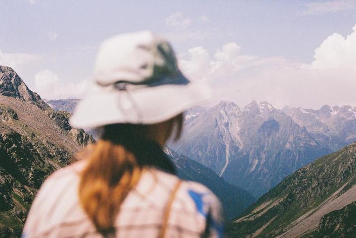 A person wearing a hat looks out at a mountain range with some snow-capped peaks.