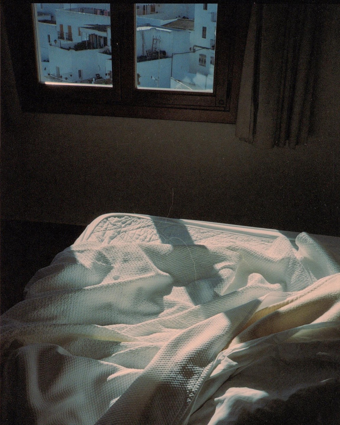 Sunlight streams through a window onto a rumpled bed, with white buildings visible outside.