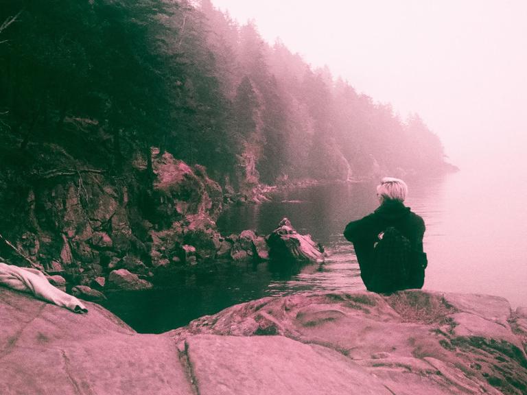 A person with light hair sits on a rock by the water, looking out at a hazy, tree-lined shore.