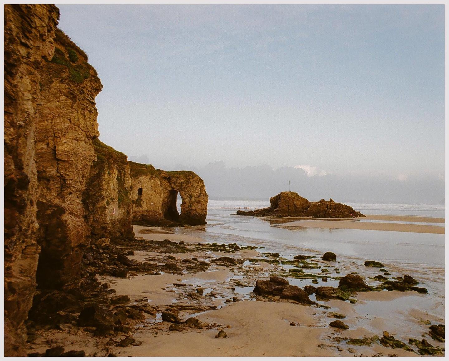 A rocky beach with a natural arch in the cliff face and a small island in the distance under a hazy sky.