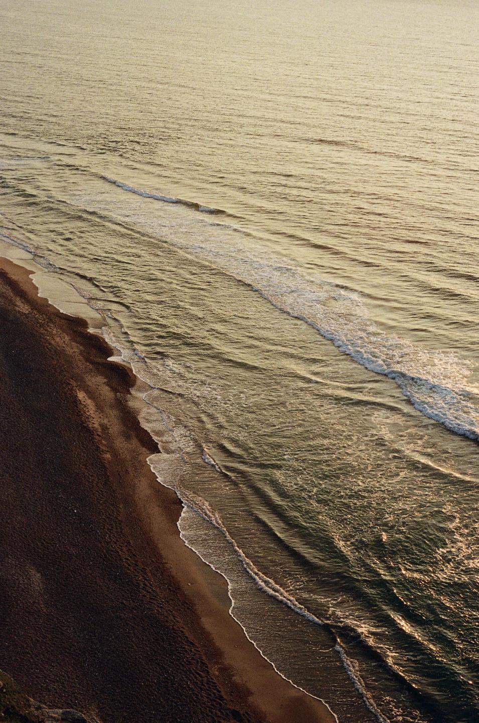 An aerial view shows waves breaking on a dark sand beach.