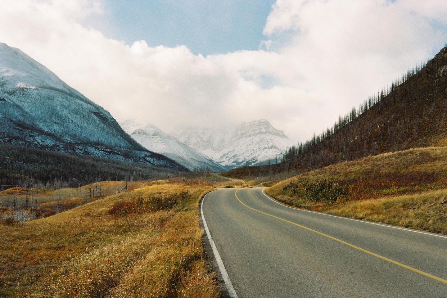 A winding road leads through a valley with snow-capped mountains and hills covered in dry grass and sparse trees under a cloudy sky.
