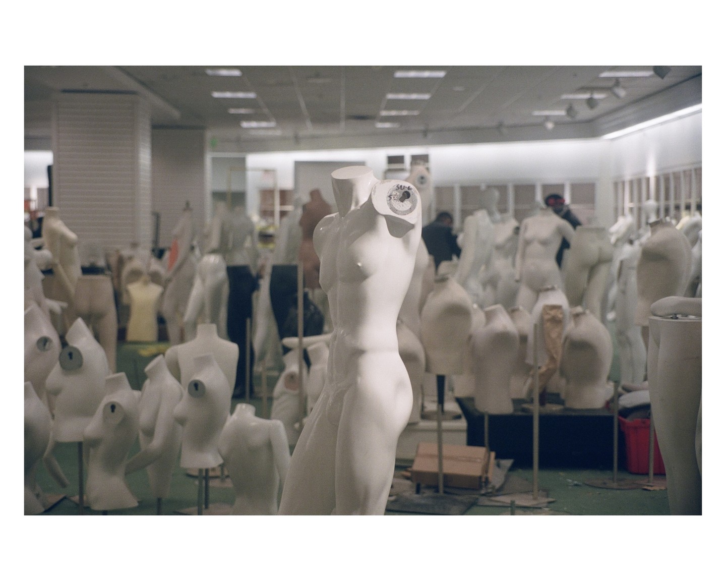 A room is filled with many white mannequins, some with limbs and some without, with a man in the background.