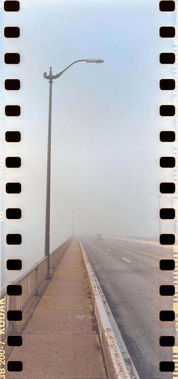 A car drives on a foggy bridge with a street light on the left.