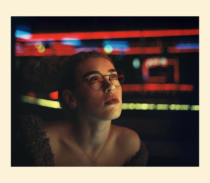 A woman with glasses and a nose ring looks up at neon lights in the dark.