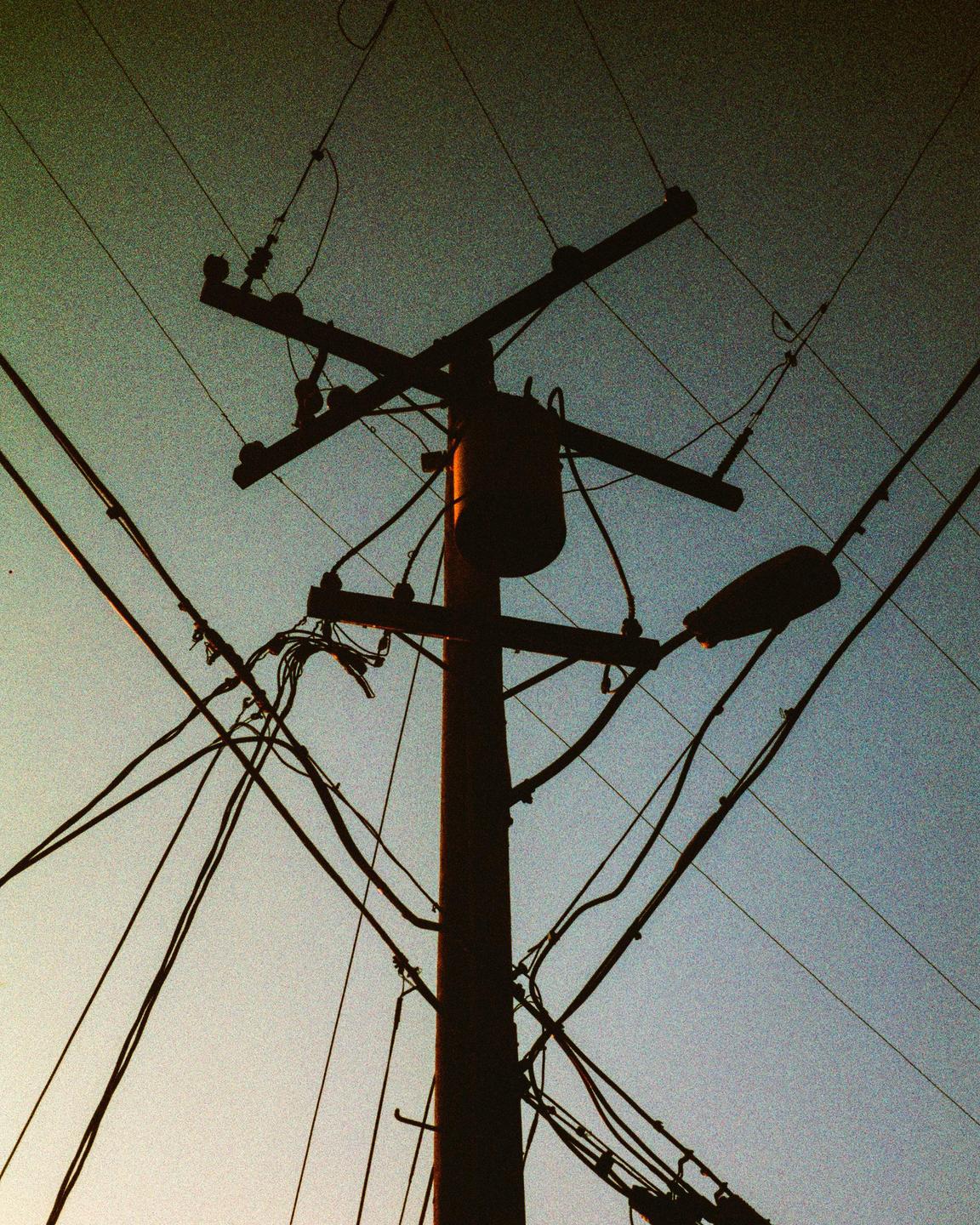 A utility pole with many wires and a street light is silhouetted against the sky.