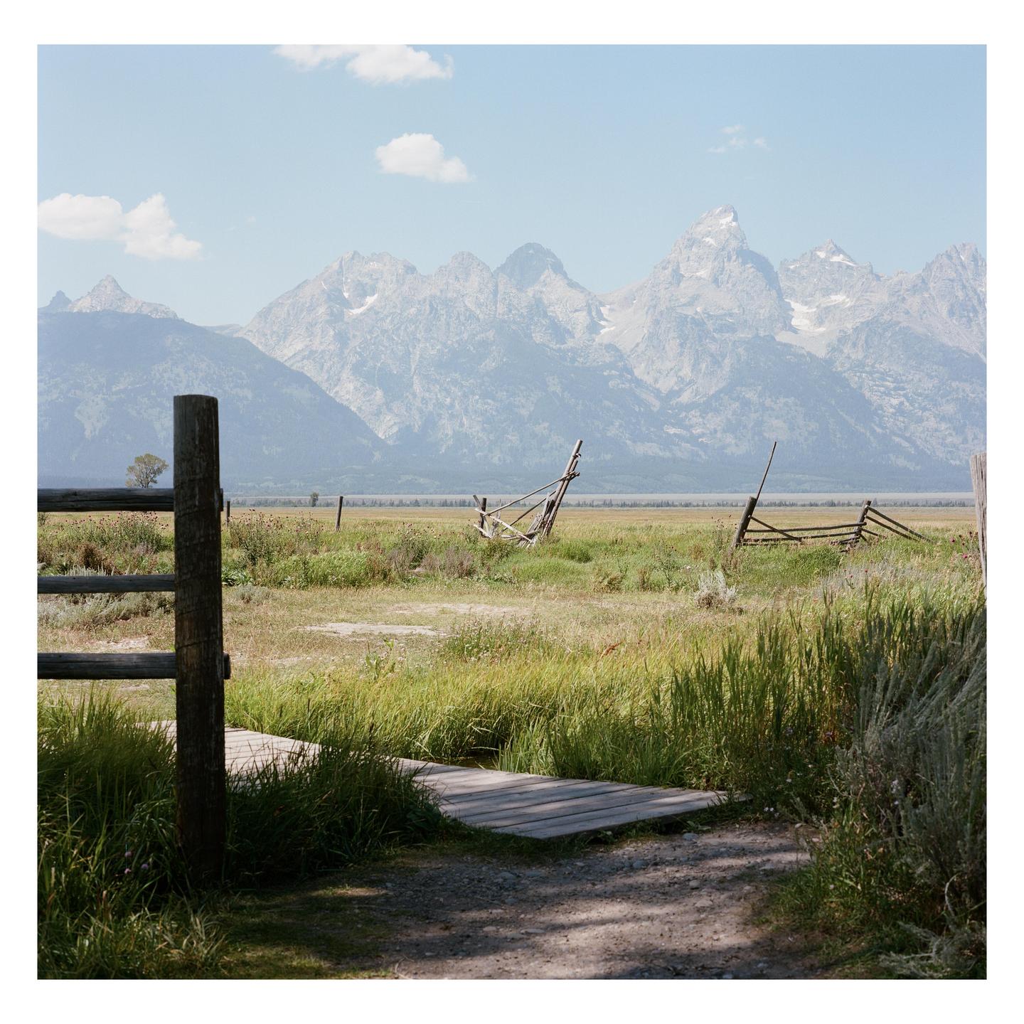 A wooden fence and boardwalk lead to a grassy field with mountains in the background.