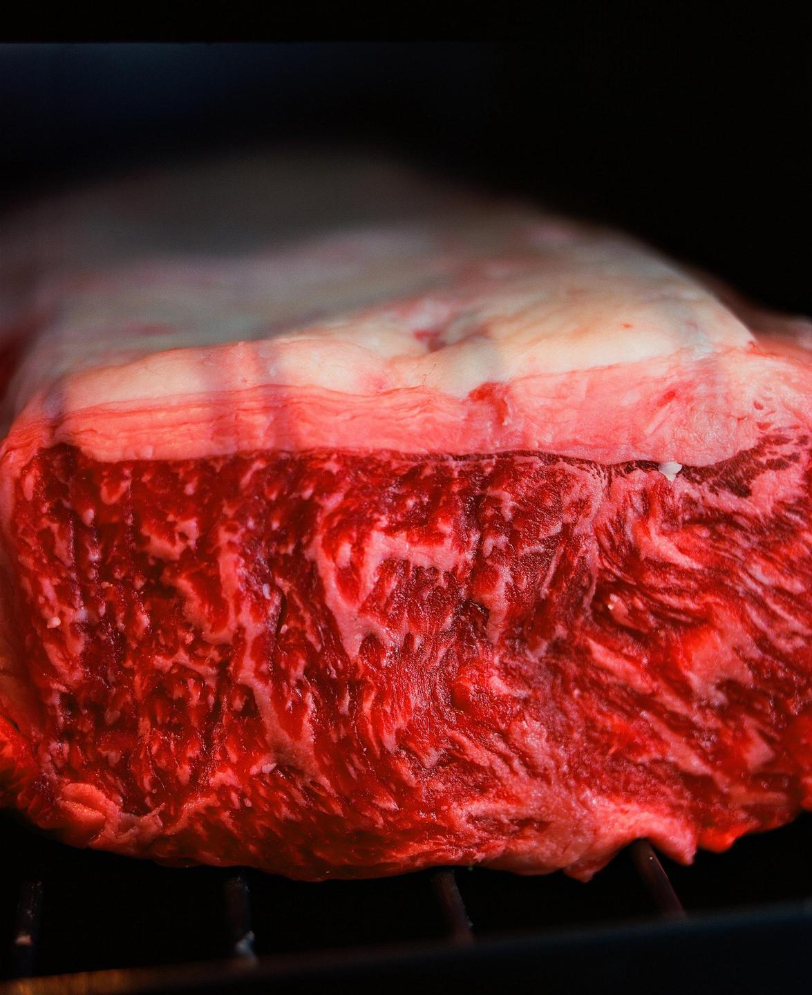 A large cut of raw meat with visible marbling rests on a grill.