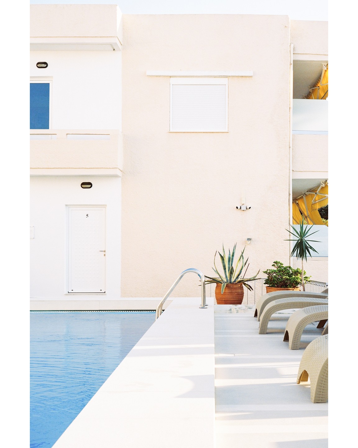 A white building with a blue window and a door with the number 5 overlooks a swimming pool with lounge chairs and potted plants.