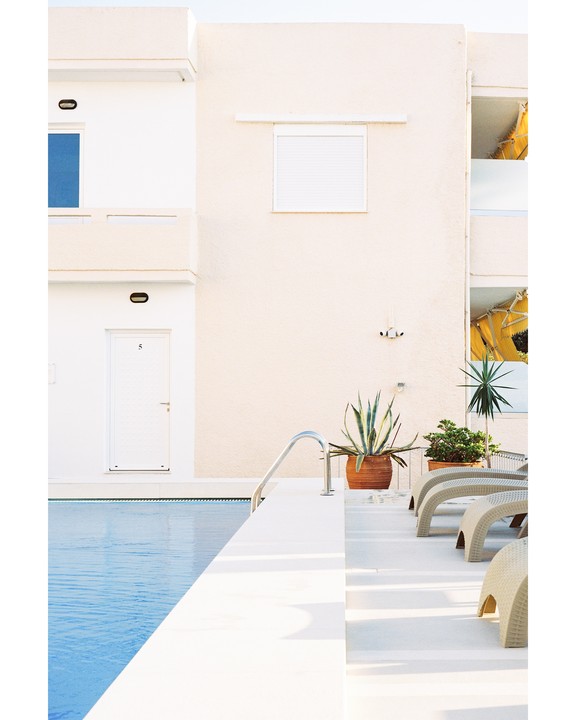 A white building with a blue window and a door with the number 5 overlooks a swimming pool with lounge chairs and potted plants.