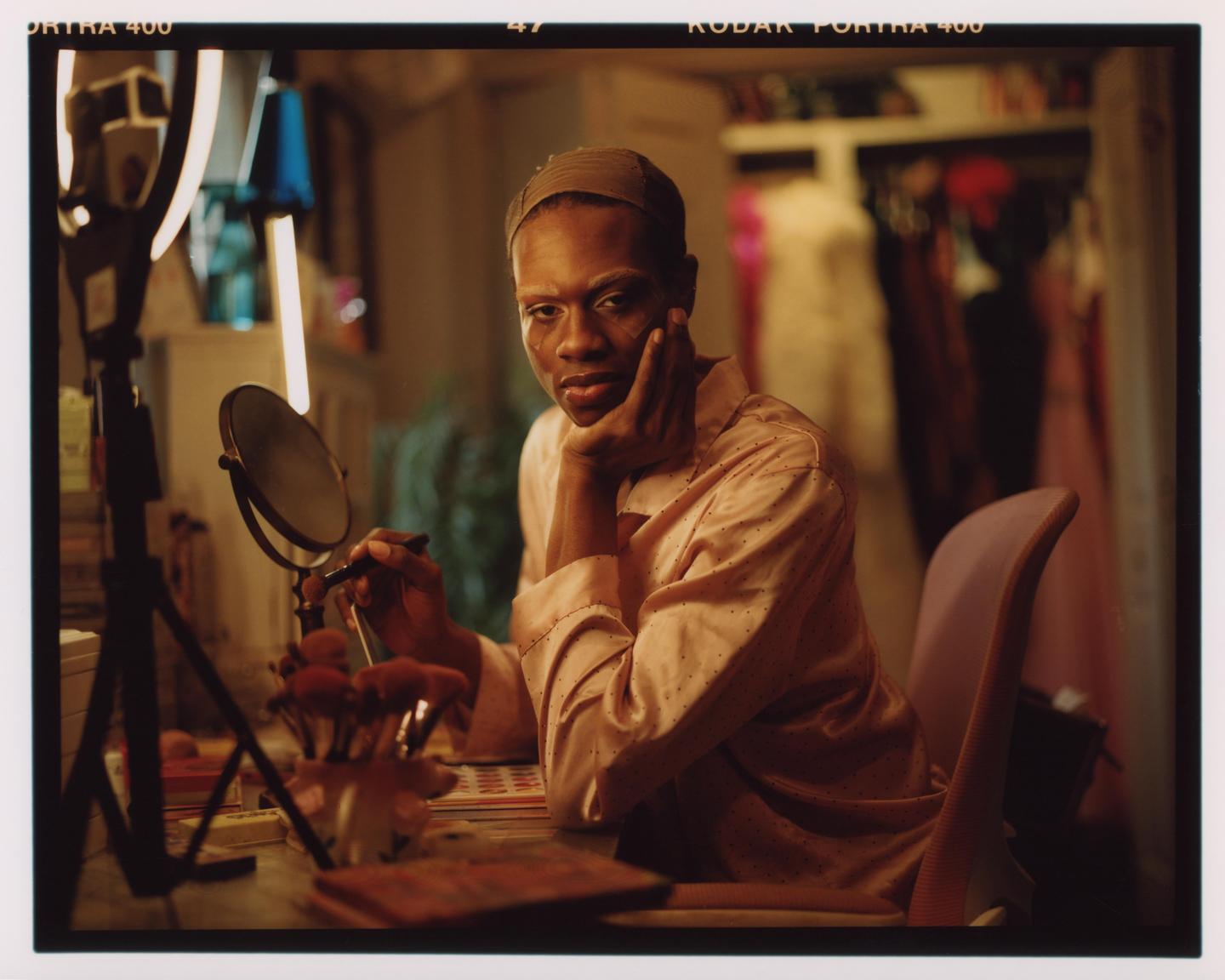 A person in a pink shirt sits at a vanity with a mirror and makeup brushes, looking towards the viewer.