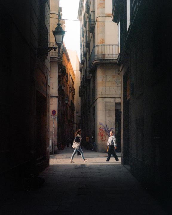 Two women walk through a narrow street with tall buildings on either side, illuminated by a patch of sunlight.