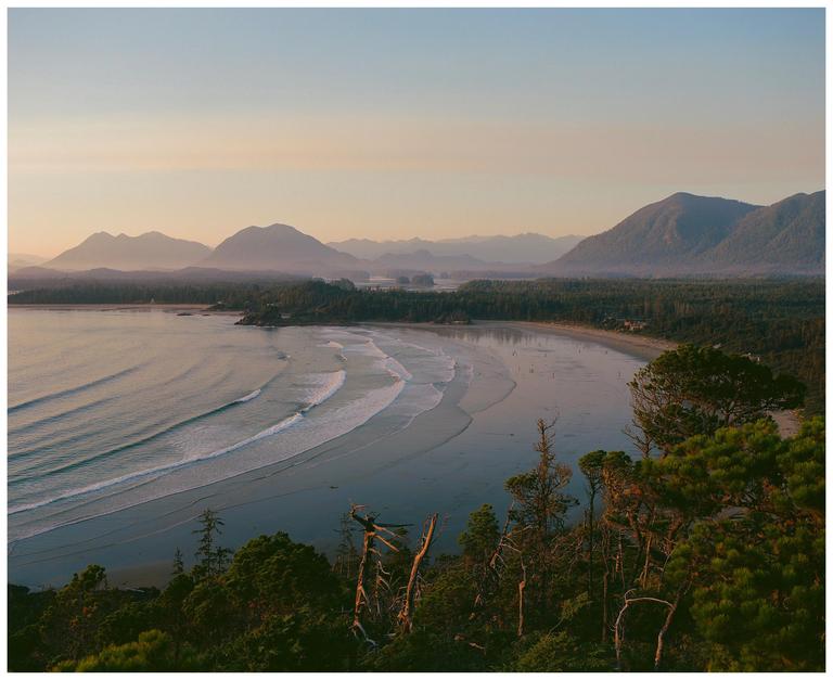 A wide shot from a high vantage point shows a beach with waves rolling in, surrounded by a forest and mountains in the distance under a hazy sky.