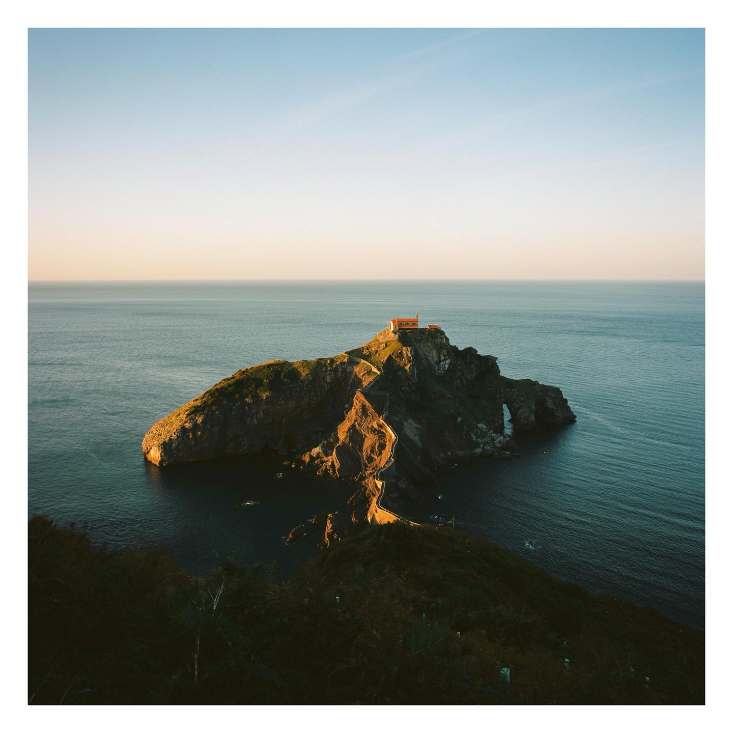 A small building sits atop a rocky island connected to the mainland by a winding path, surrounded by the sea under a clear sky.