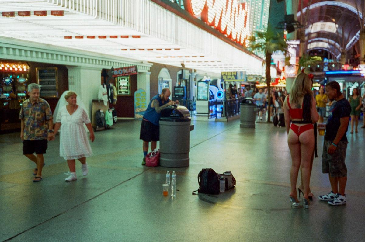 A woman in a white dress and a man in a patterned shirt walk hand-in-hand past a man looking into a trash can and a woman in a red bikini talking to a man.