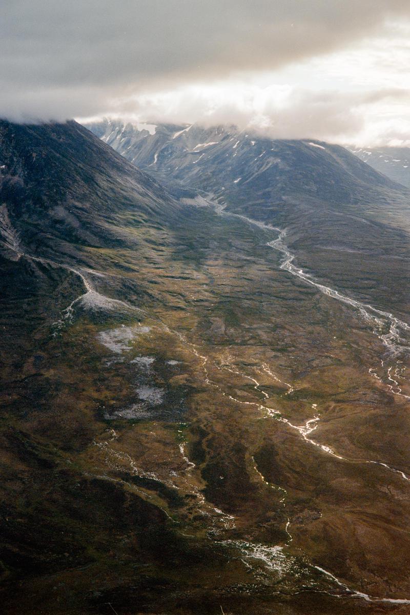 A winding river flows through a valley surrounded by mountains with clouds obscuring their peaks.