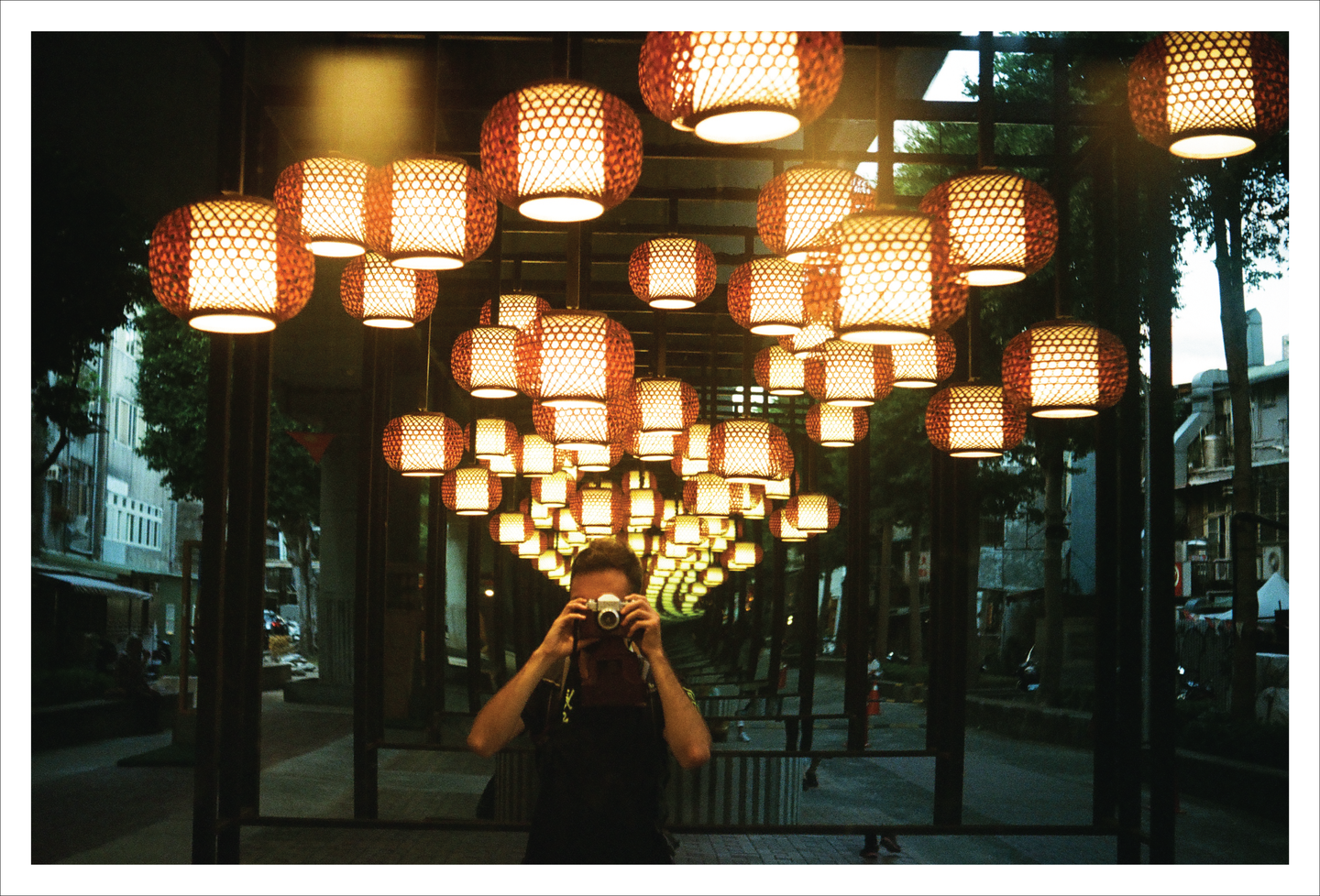 A man holds a camera to his face, reflected in a tunnel of illuminated lanterns.