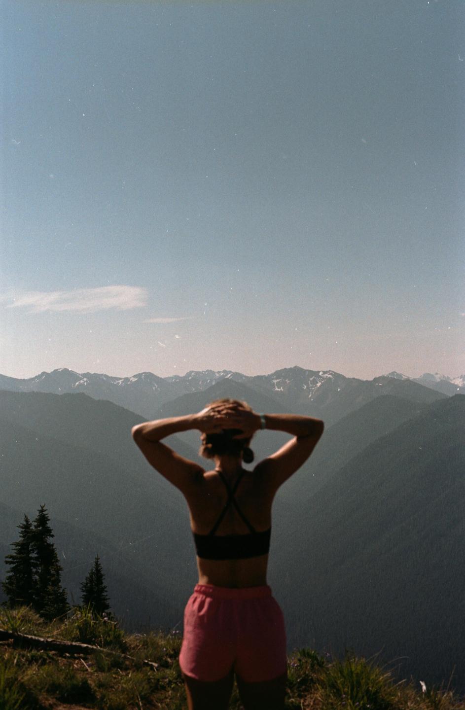 A woman stands with her back to the camera, looking out at a mountain range under a clear sky.
