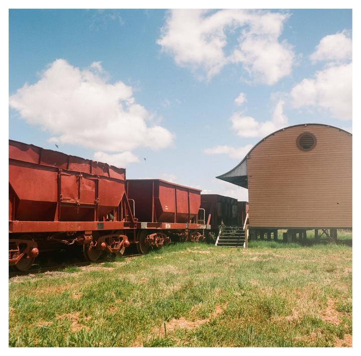 Red train cars sit on tracks next to a wooden building with a curved roof under a blue sky with clouds.