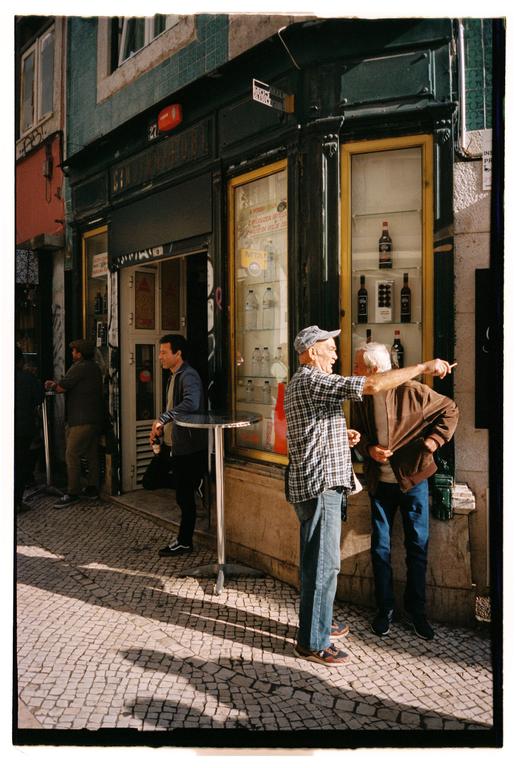 Two men stand on a cobblestone street in front of a shop, one pointing into the distance.