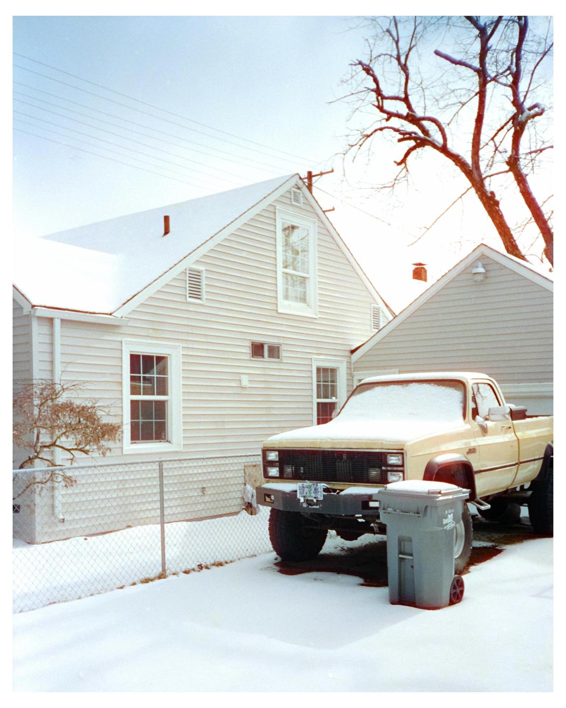 A light-colored pickup truck covered in snow is parked next to a house with a snow-covered roof.