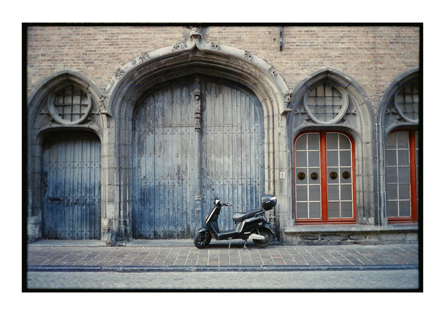A black scooter is parked on a cobblestone street in front of a brick building with large wooden doors and red-framed windows.