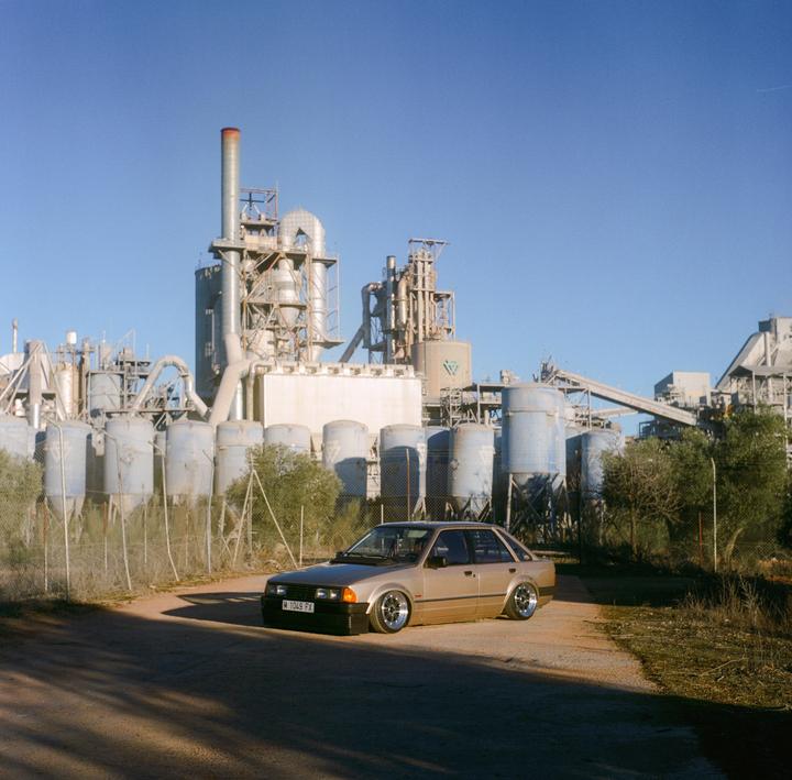 A gold car is parked on a dirt road in front of an industrial plant under a clear blue sky.