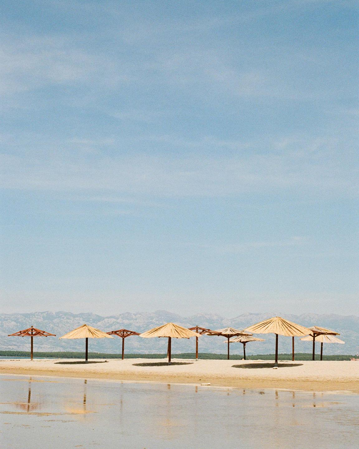 A row of straw beach umbrellas stands on a sandy beach with mountains in the background.
