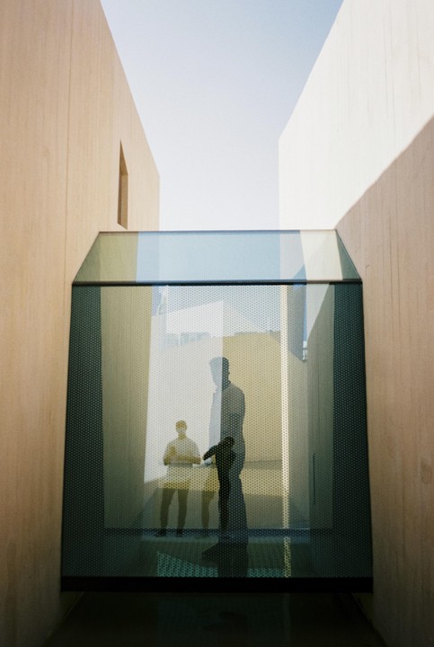 Two men are visible through a perforated glass structure in a modern building with tall, plain walls.