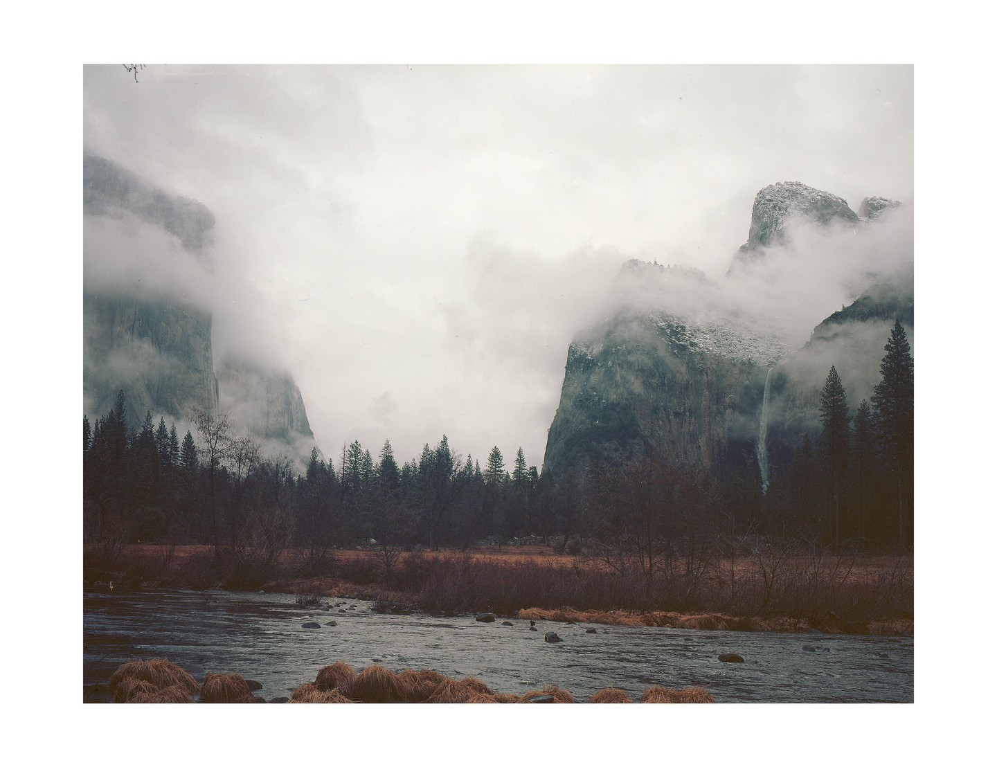 A river flows through a valley with tall mountains shrouded in fog and evergreen trees.