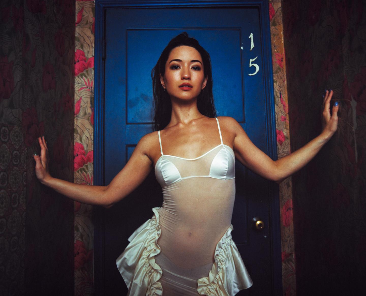 A woman in a sheer white outfit stands in front of a blue door with the number 15 on it, next to a wall with floral wallpaper.
