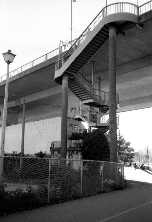A spiral staircase descends from an overpass next to a chain link fence and a street light.