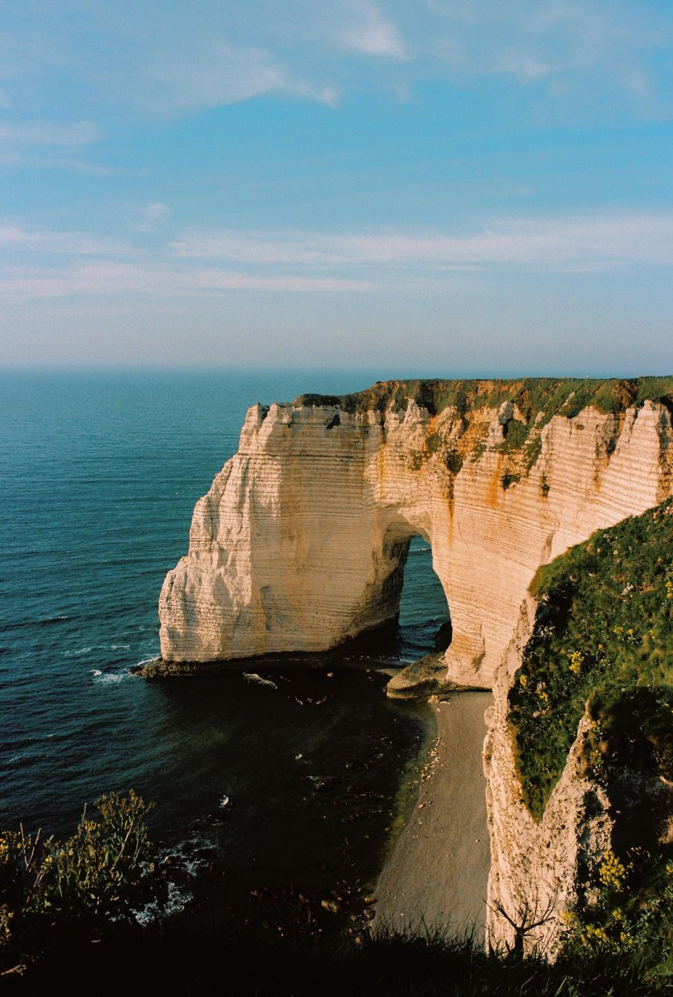 A large rock formation with an archway stands in the ocean next to a cliff covered in green foliage.