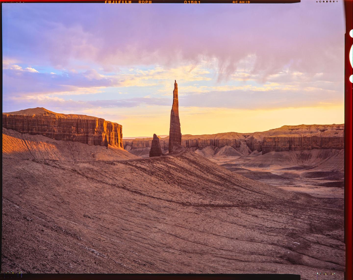 A tall, thin rock formation stands prominently in a vast desert landscape under a colorful sky at sunset.