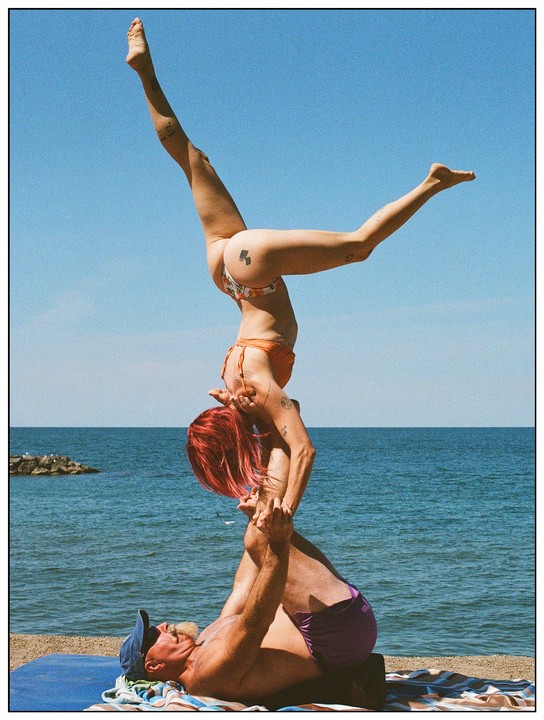 A man lies on his back on a beach towel, supporting a woman with red hair who is balancing on his feet with her legs extended upwards.