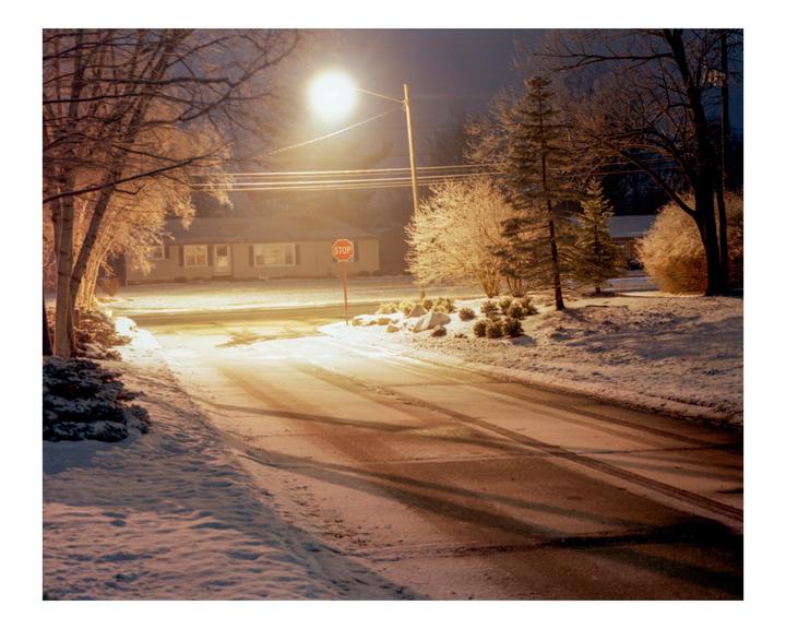 A snow-covered street with a stop sign is illuminated by a street light at night.
