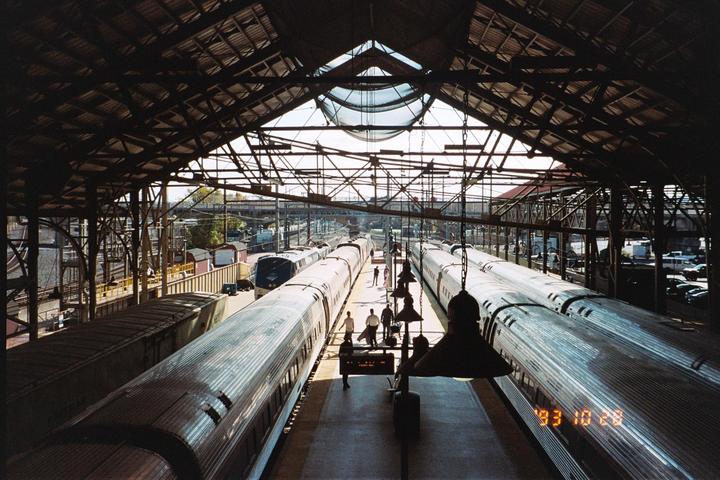 Two trains are parked at a station with people walking on the platform under a large, arched roof.